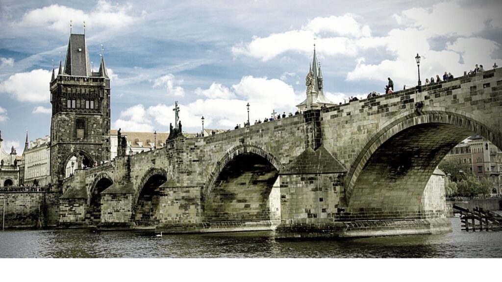 Best cities to visit in Europe: Panoramic view of Charles Bridge in Prague with the Old Town Bridge Tower on the left and Prague Castle in the distance under a partly cloudy sky.